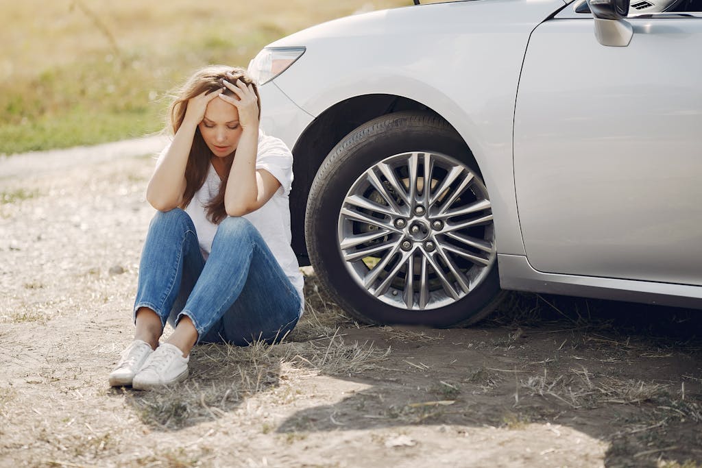 unexpected expense frustrated female driver in white t shirt and jeans sitting on ground near damaged car with hands on head during car travel in sunny summer day